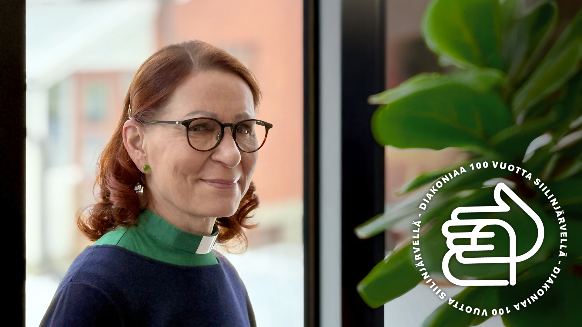 Liisa Tiilikainen ikkunan edessä, vieressä viherkasvi, jonka päällä kuvassa on diakonian juhlavuoden logo