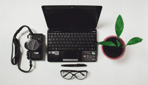 kuvassa tietokone, kamera, silmälasit, kynä ja viherkasvi