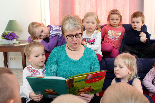 Nainen lukee lapsille kirjaa. Nainen istuu lattialla. Lapset ovat hänen ympärillään.