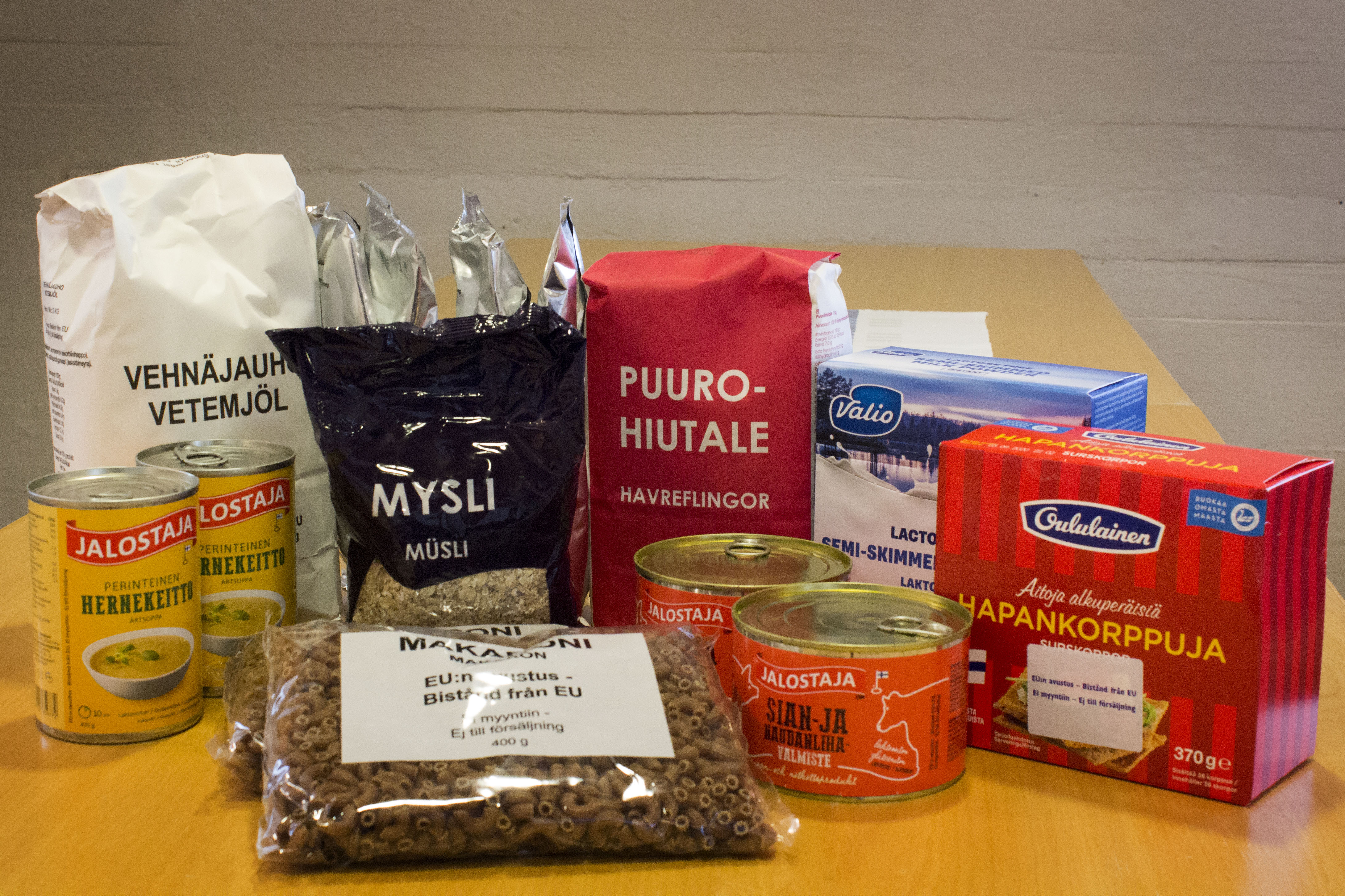 EU-ruokapaketin tuotepakkauksia pöydällä: mm. makaronia, puurohiutaleita, säilykepurkkeja ja hapankorppuja.