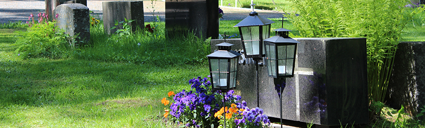 Hautakivi, lyhtyjä ja kukkia kesäisellä hautausmaalla.